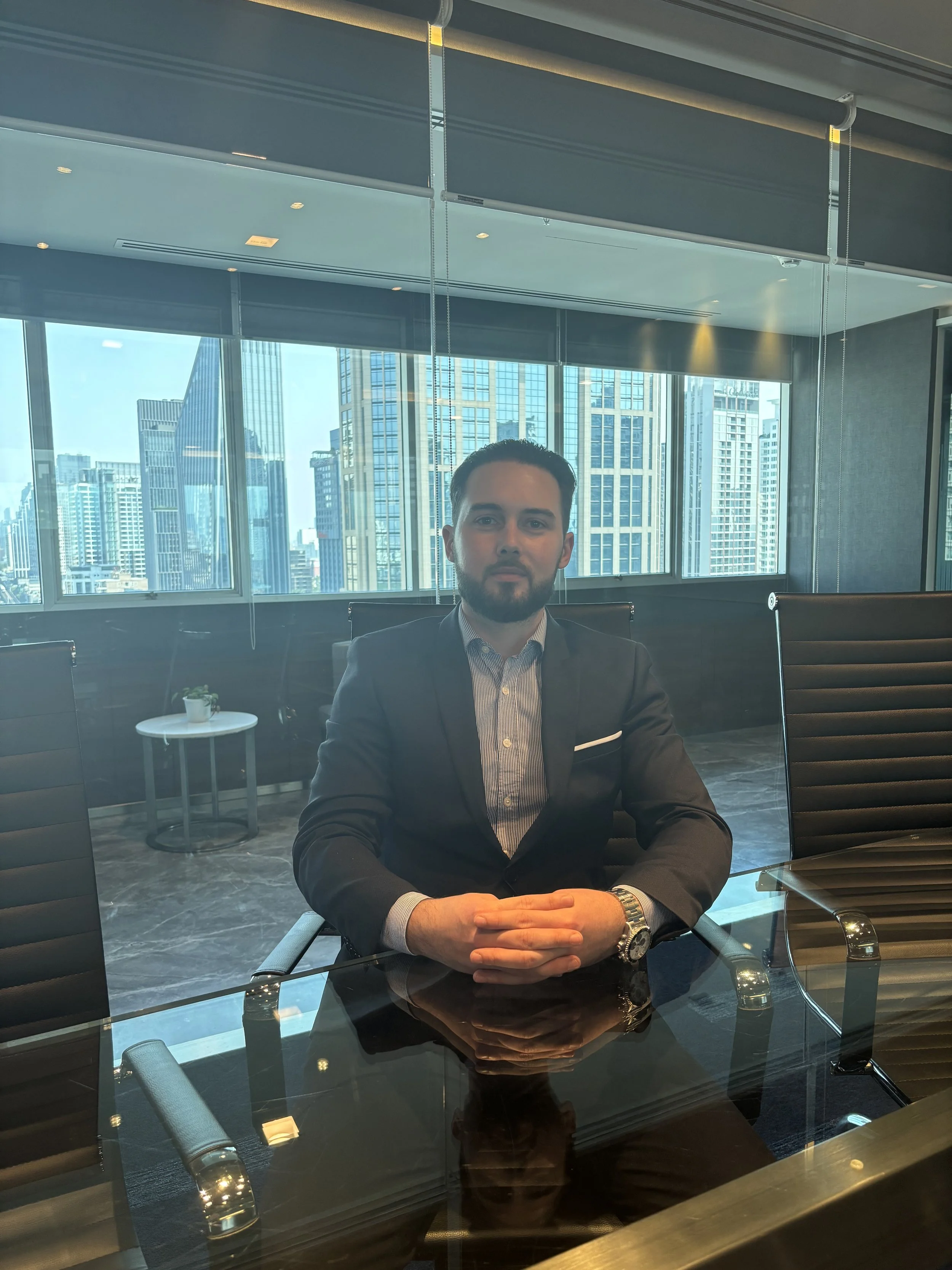 Un homme d'affaires habillé en costume assis à une table de conférence dans un bureau moderne avec de grandes fenêtres offrant une vue sur des gratte-ciel urbains.