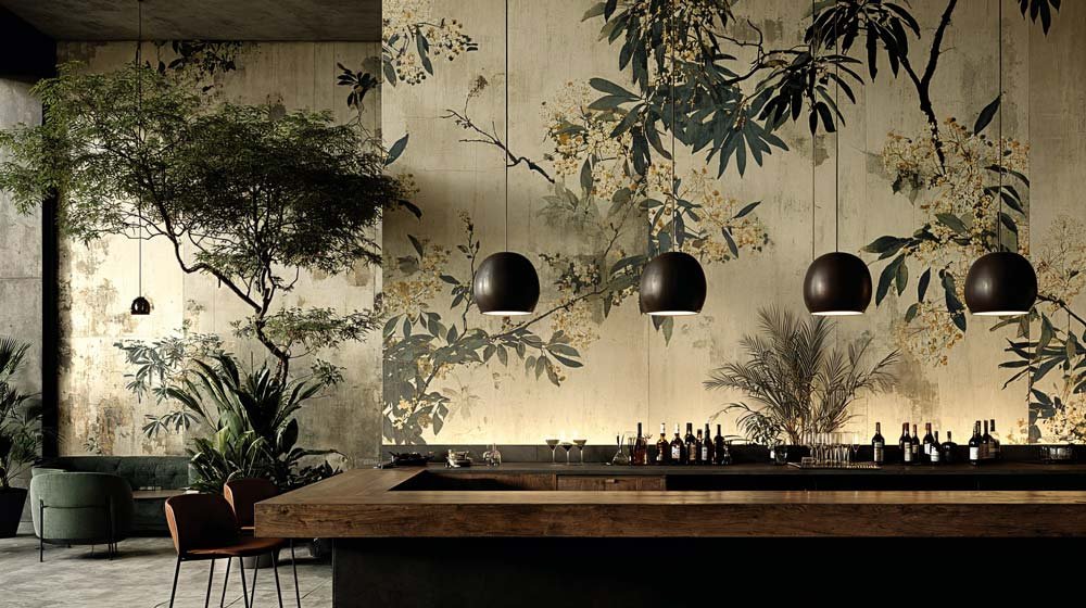 Ein stilvoller Barbereich mit dunklen Hängelampen, grünen Pflanzen und einem Wandmural mit Blumen- und Baummustern, sowie einem langen Holzbar mit Flaschen und Gläsern.