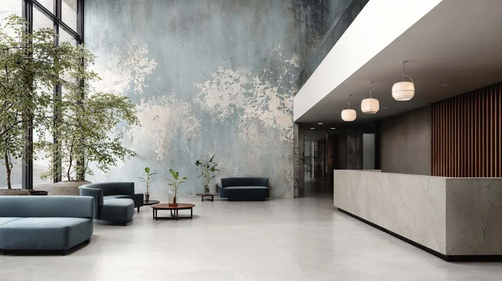 Modernes Hotel-Lobby mit grauer Wand, großen Fenstern, grünen Pflanzen, schwarzen und blauen Sitzmöbeln und weißer Empfangstheke.