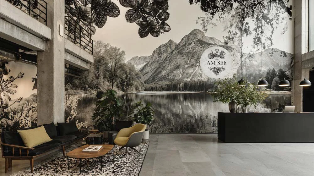 Ein modernes Hotel-Lobby mit großer Wandtapete eines Sees und Berge, einem schwarzen Empfangstresen, Pflanzen, Sitzmöbeln und Beleuchtung.