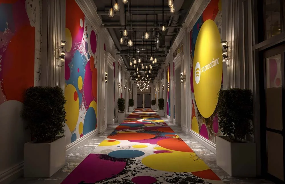 Ein farbiger, moderner Flur mit bunten, großen Wanddekorationen und Teppichmustern, Pflanzen in weißen Pflanzkübeln und einer Deckenbeleuchtung mit mehreren Glühbirnen.