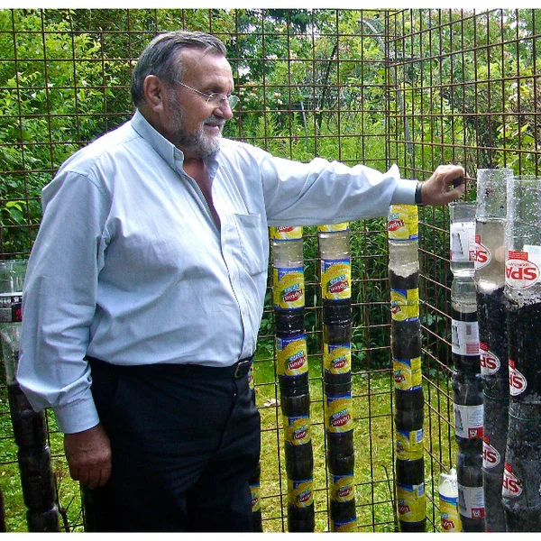 A man with gray hair and beard, wearing a white shirt and black pants, stands outdoors inside a metal enclosure with trees and greenery in the background. He is smiling and holding a plastic bottle filled with dark liquid, looking at several vertical columns of similar bottles attached to the enclosure.