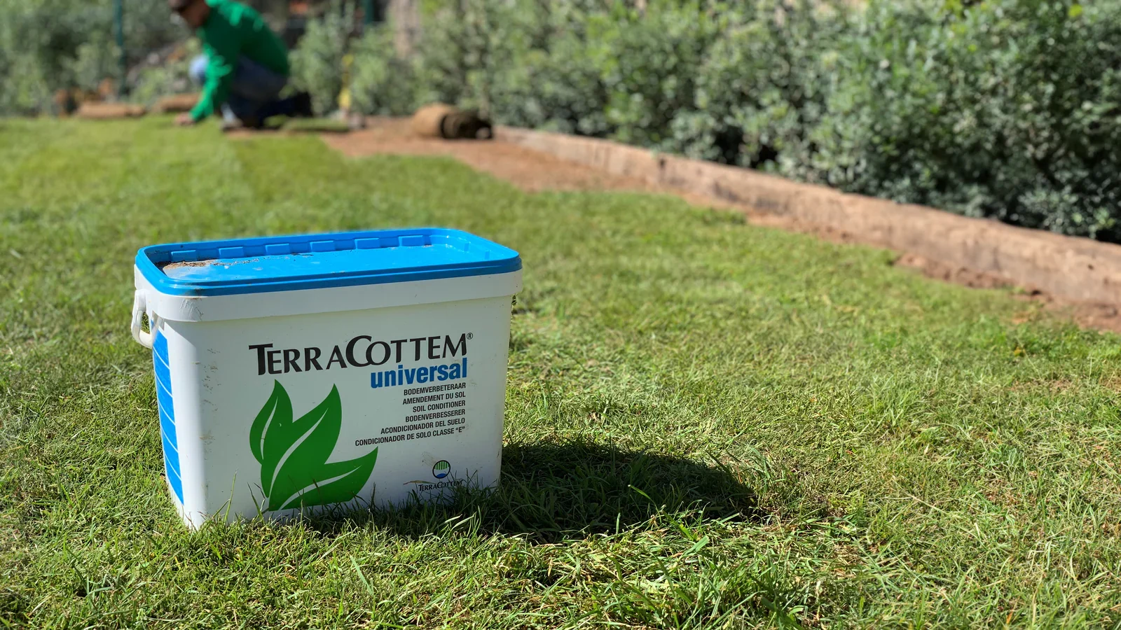 A bucket of TerraCottem soil conditioner on a lawn with a gardener working in the background.