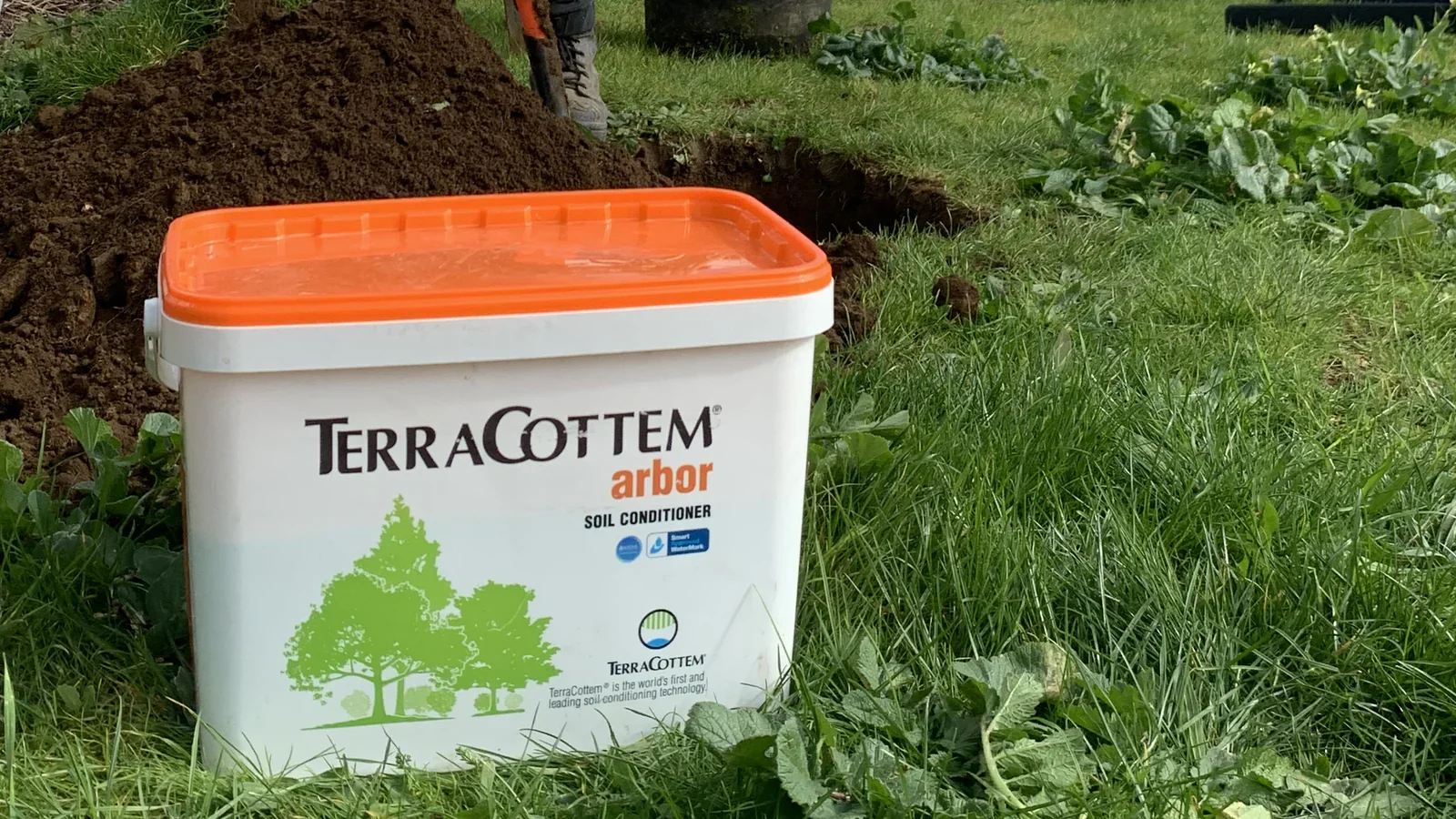 A white container of TerraCottem arbor soil conditioner with an orange lid, placed on the ground next to a garden bed with freshly turned soil and green plants.