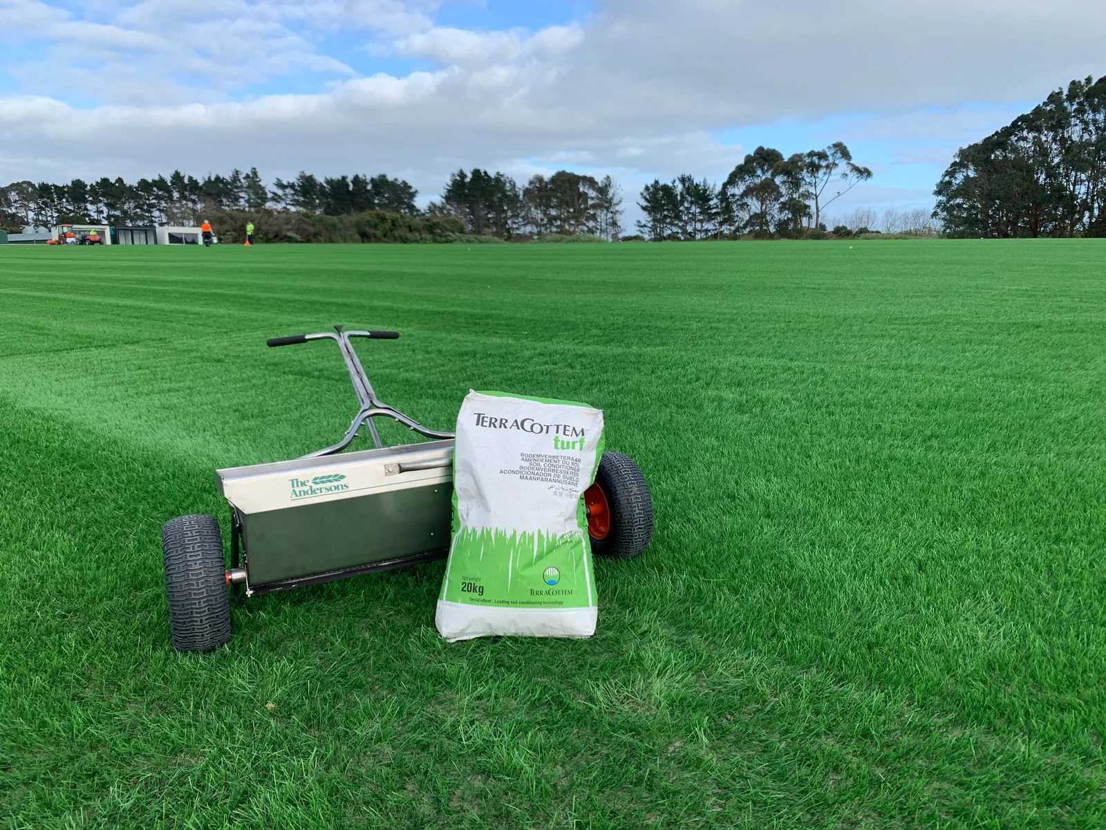 A turf management machine and a bag of turf soil on a well-maintained green sports field, with trees and a partly cloudy sky in the background.