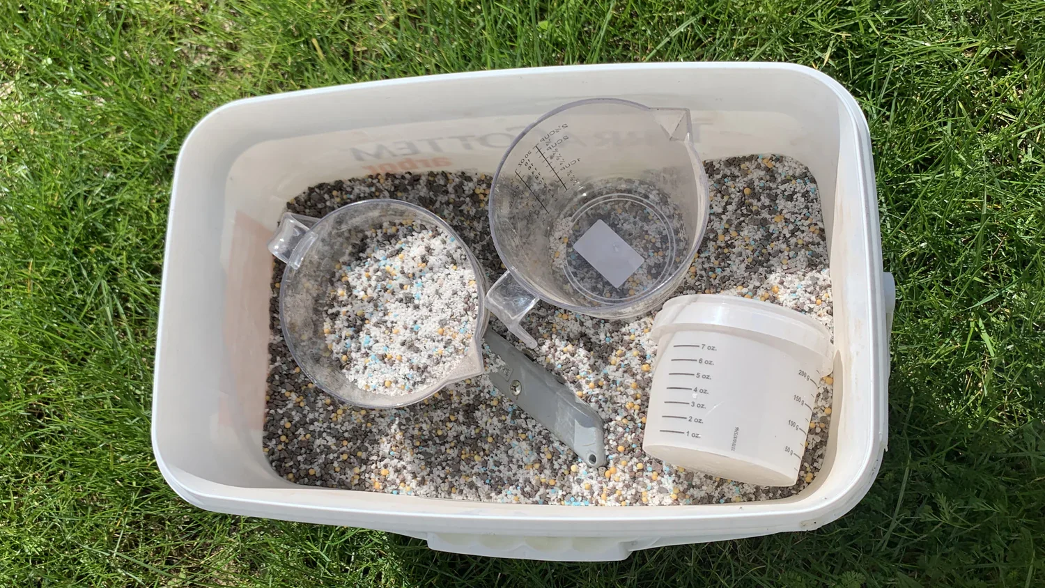 Container filled with a mixture of small colorful rocks and sand, measuring cups and a pair of scissors inside, located outdoors on green grass.