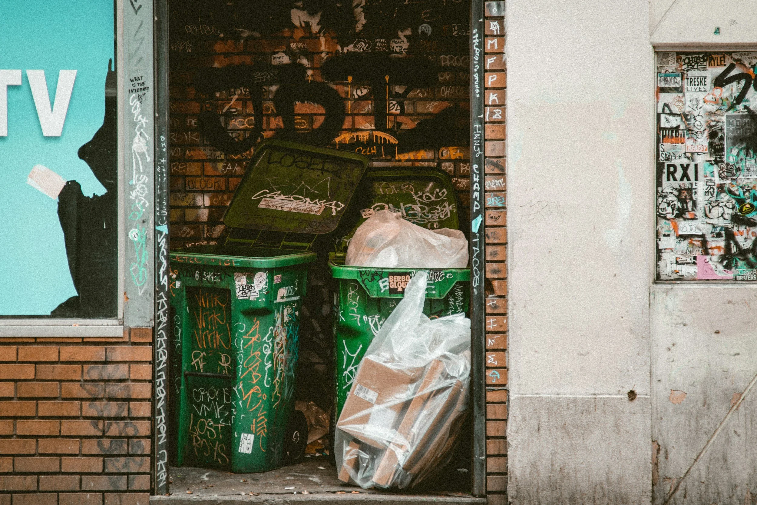 Gatumiljö med två gröna sopkärl fyllda med skräp och en plastpåse, omgärdade av graffiti och väggar.