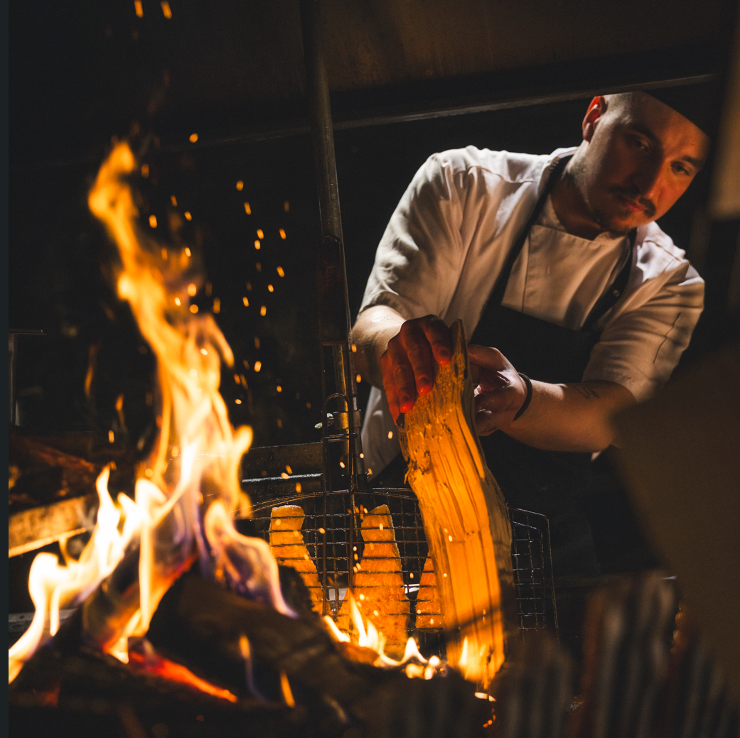 A chef in a white uniform and black apron cooking over an open flame, adding a large piece of wood to the fire in a professional kitchen.