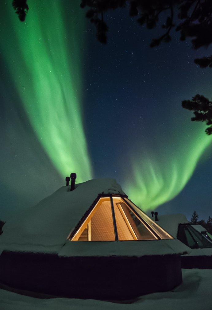 A snowy A-frame cabin at night illuminated from inside, with vibrant northern lights and stars in the sky above.