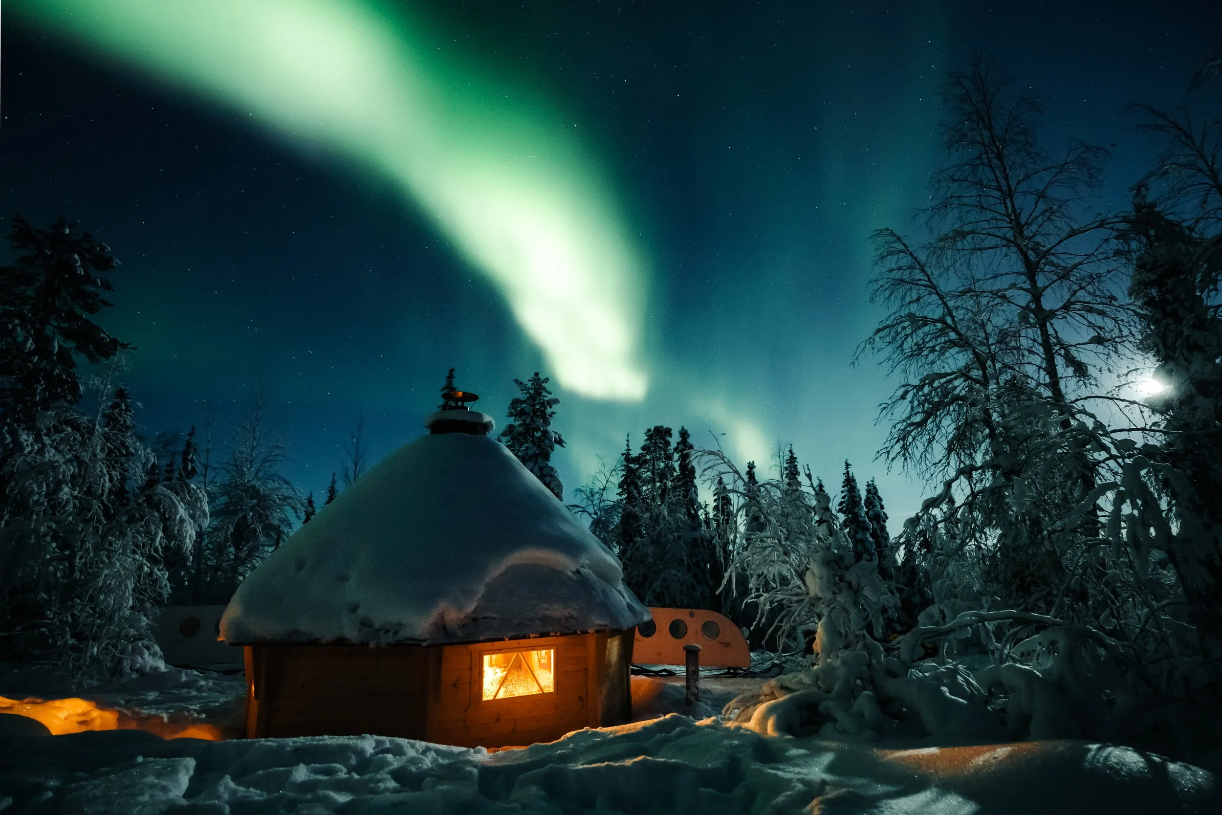 A snowy winter landscape at night featuring the Northern Lights in the sky. A cozy wooden cabin with a lit window is visible, surrounded by snow-covered trees. The northern lights display a greenish glow across the starry night sky.