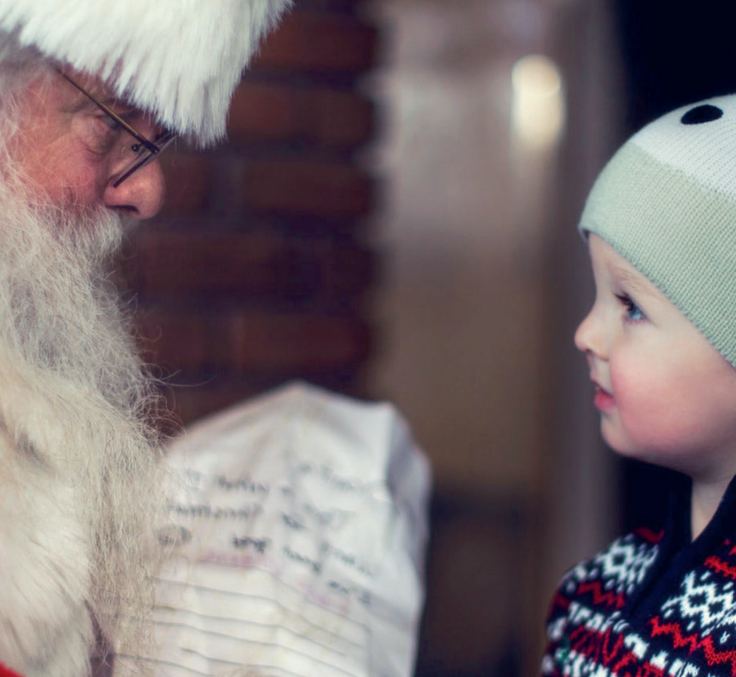 A young boy in a winter hat and sweater looking at Santa Claus, who is dressed in a traditional red suit with white fur trim, with a blurred indoor background.