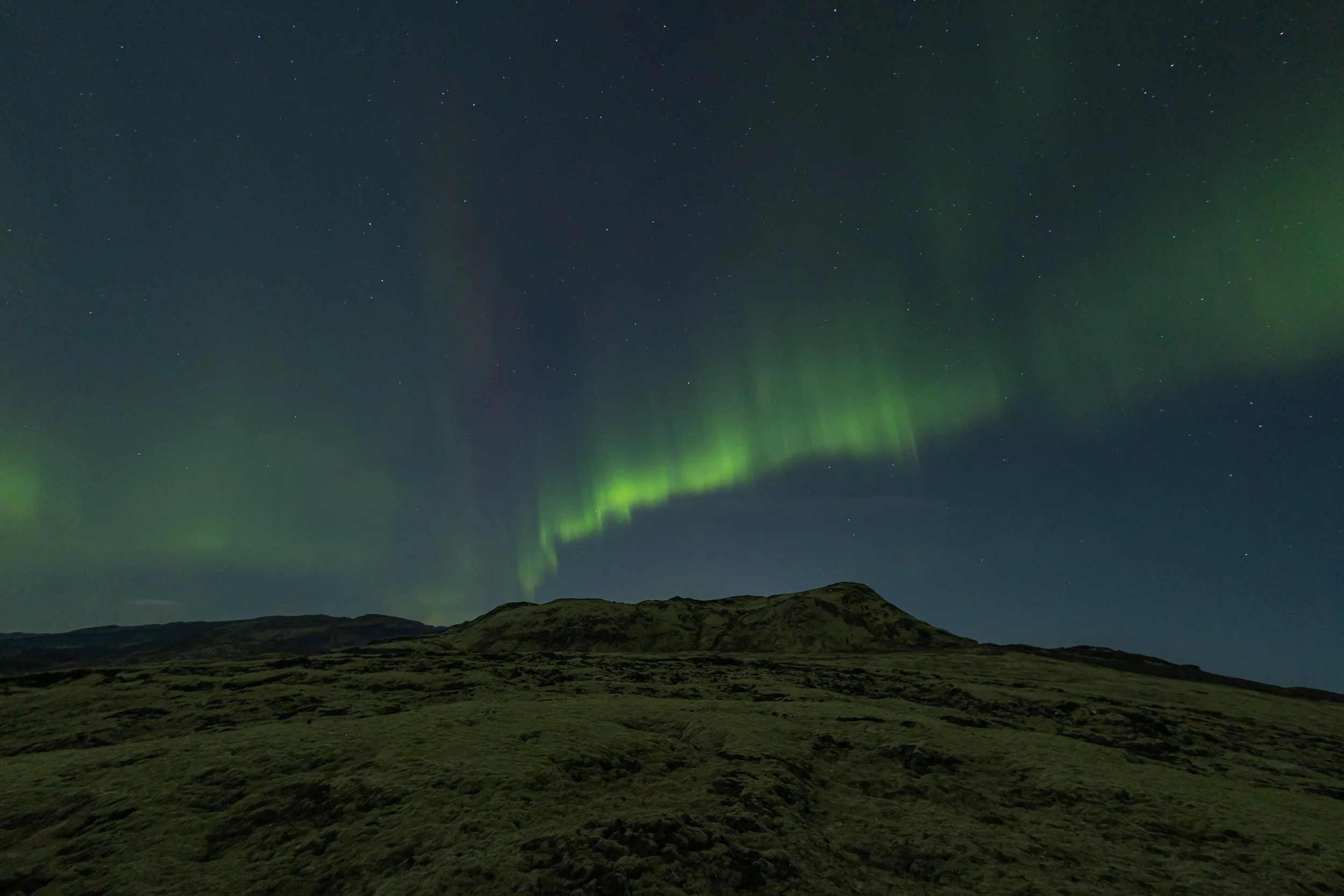 Nighttime landscape with green aurora borealis lighting up the sky above rolling hills.