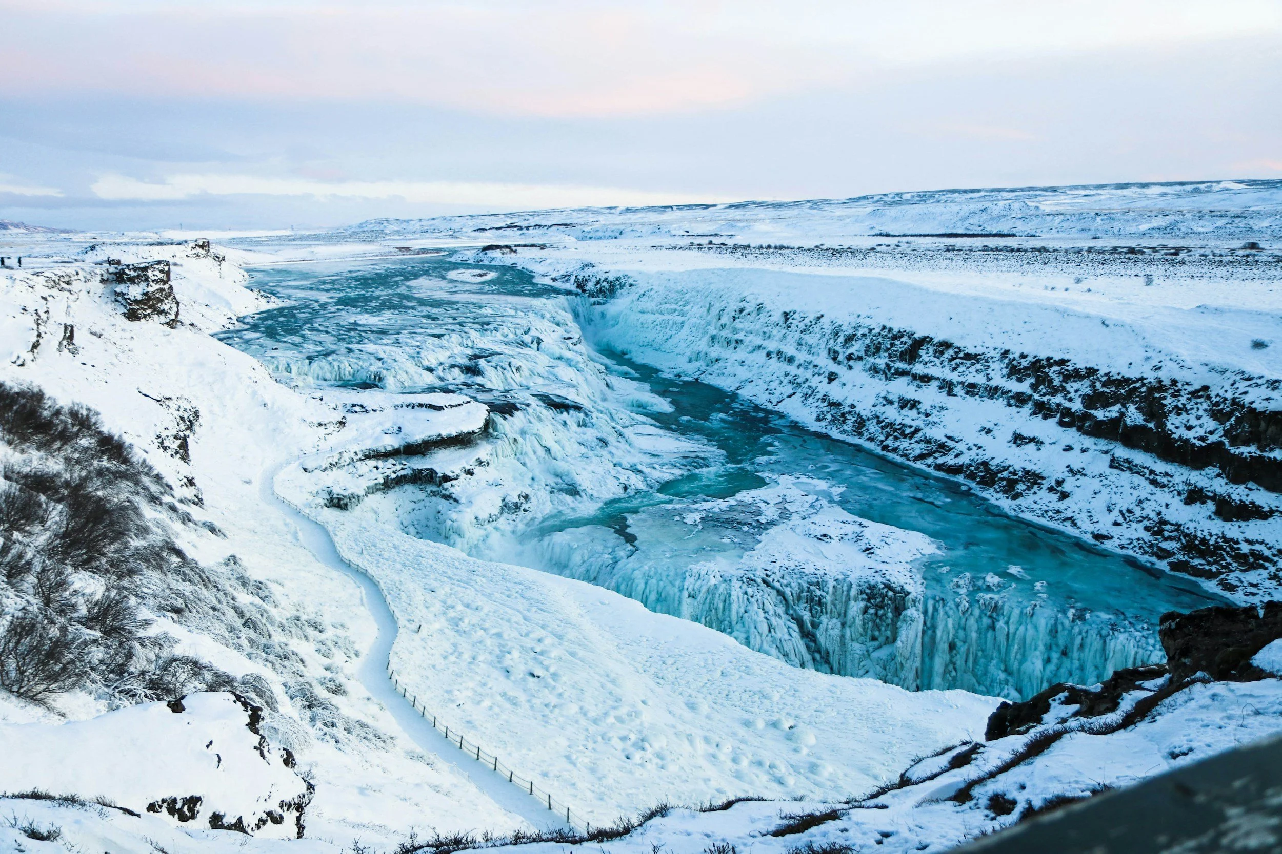 A snowy landscape featuring a large waterfall or fjord with ice and snow-covered cliffs, and a partially frozen river flowing through the area under a cloudy sky.