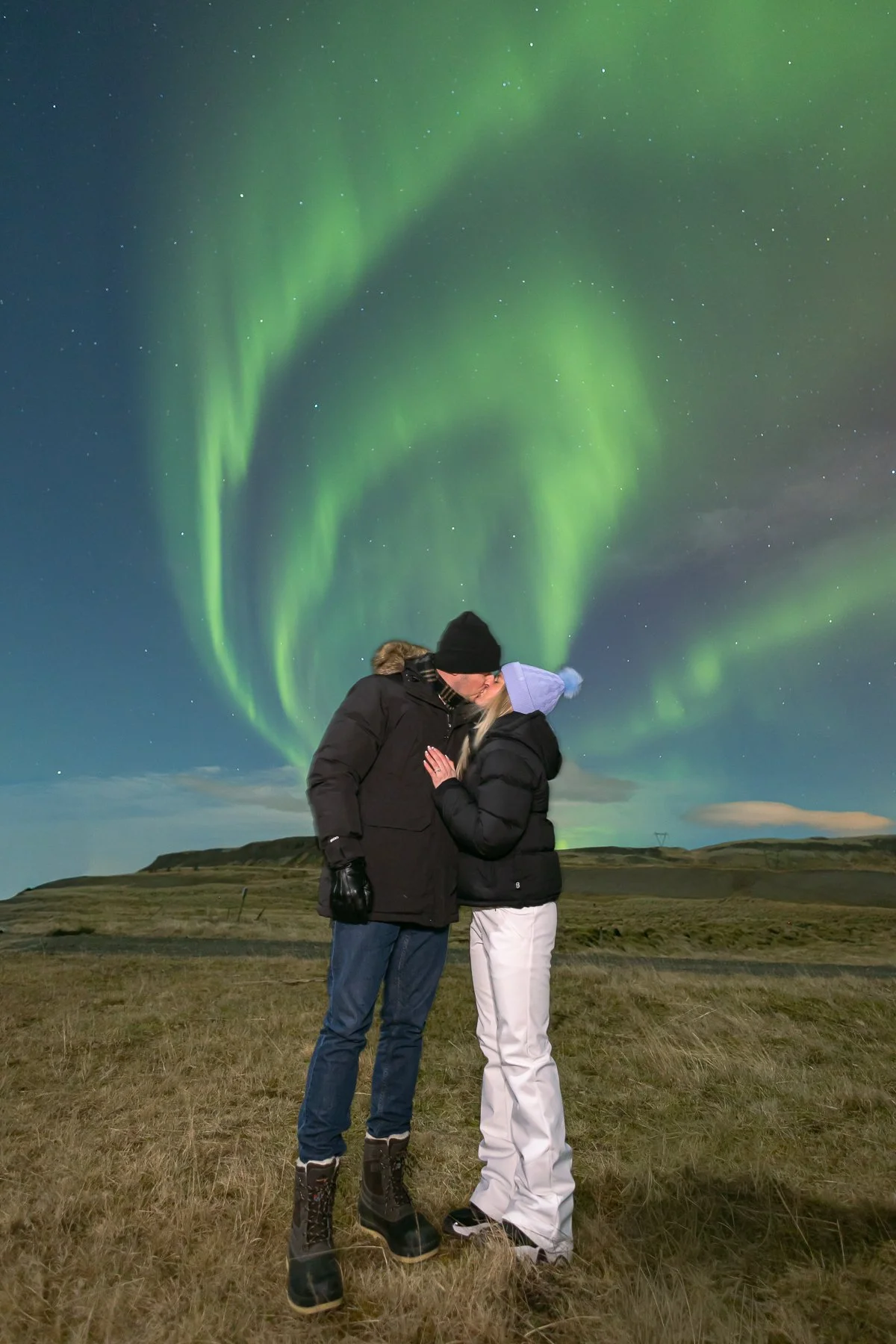 Two people kissing outdoors at night, with the Northern Lights in the sky above them.