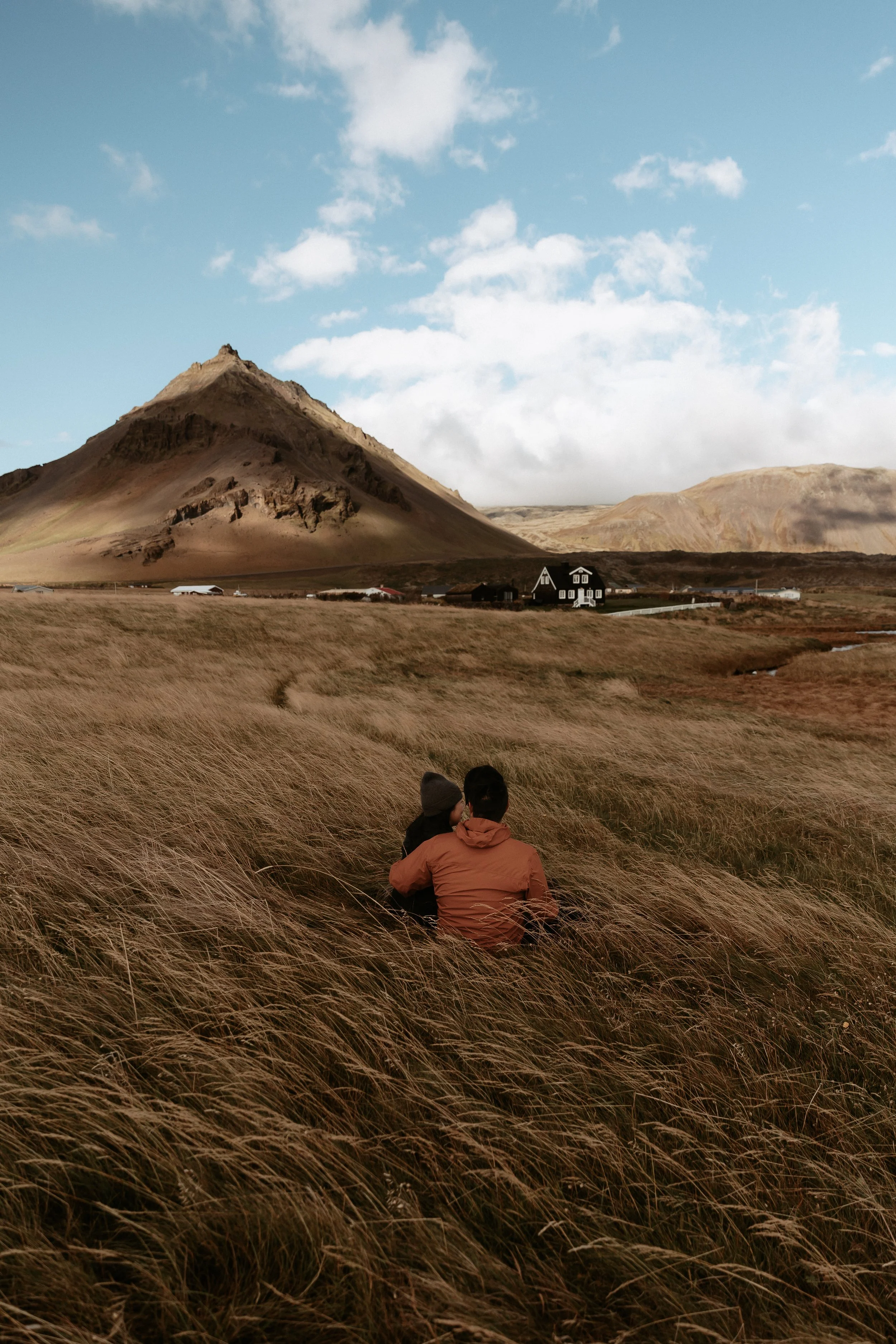 A couple sitting in tall grass overlooking a mountain in a rural area.