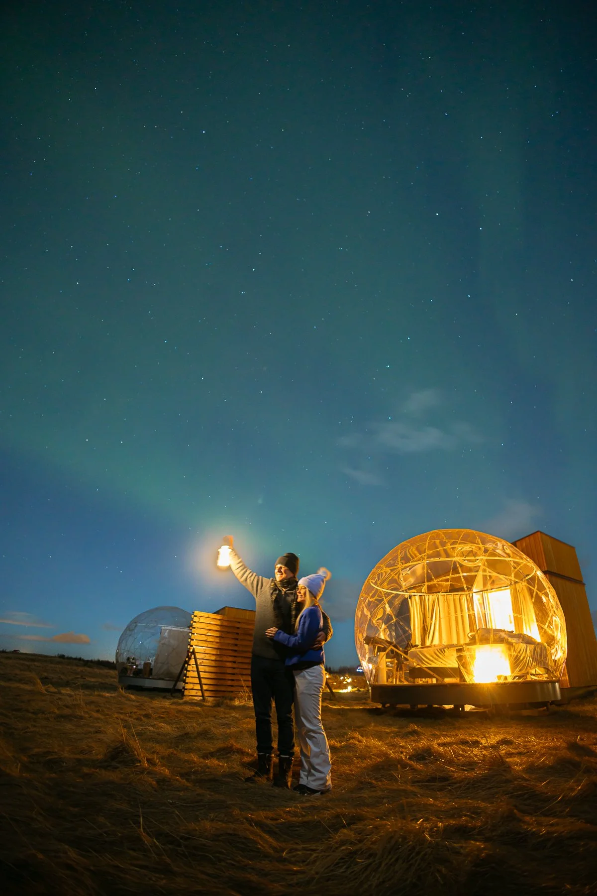 Two people stand outside at night taking a selfie with a lantern, in front of two illuminated transparent geodesic dome structures under a starry sky.