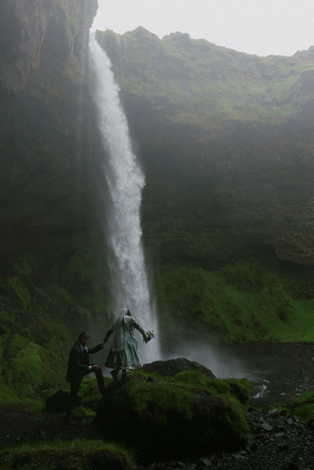 Couple on a private day tour in Iceland admiring Kvernufoss waterfall standing at the base of a waterfall in a lush, green landscape. T