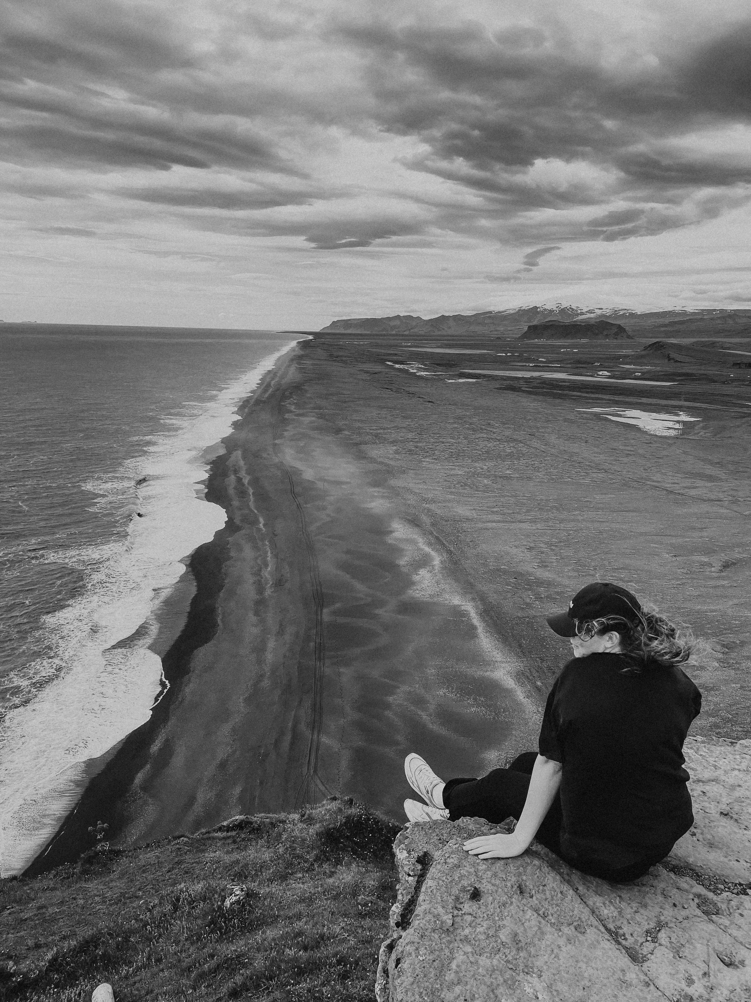 A person sitting on a rocky ledge overlooking a coastline with a long beach and ocean waves, with mountains and cloudy sky in the distance.
