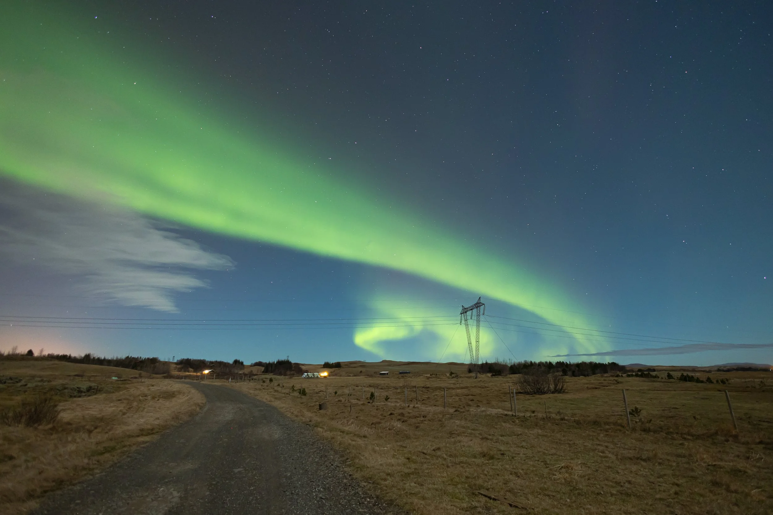 Nighttime landscape with a dirt road, open field, power line tower, and vibrant green northern lights across the starry sky.