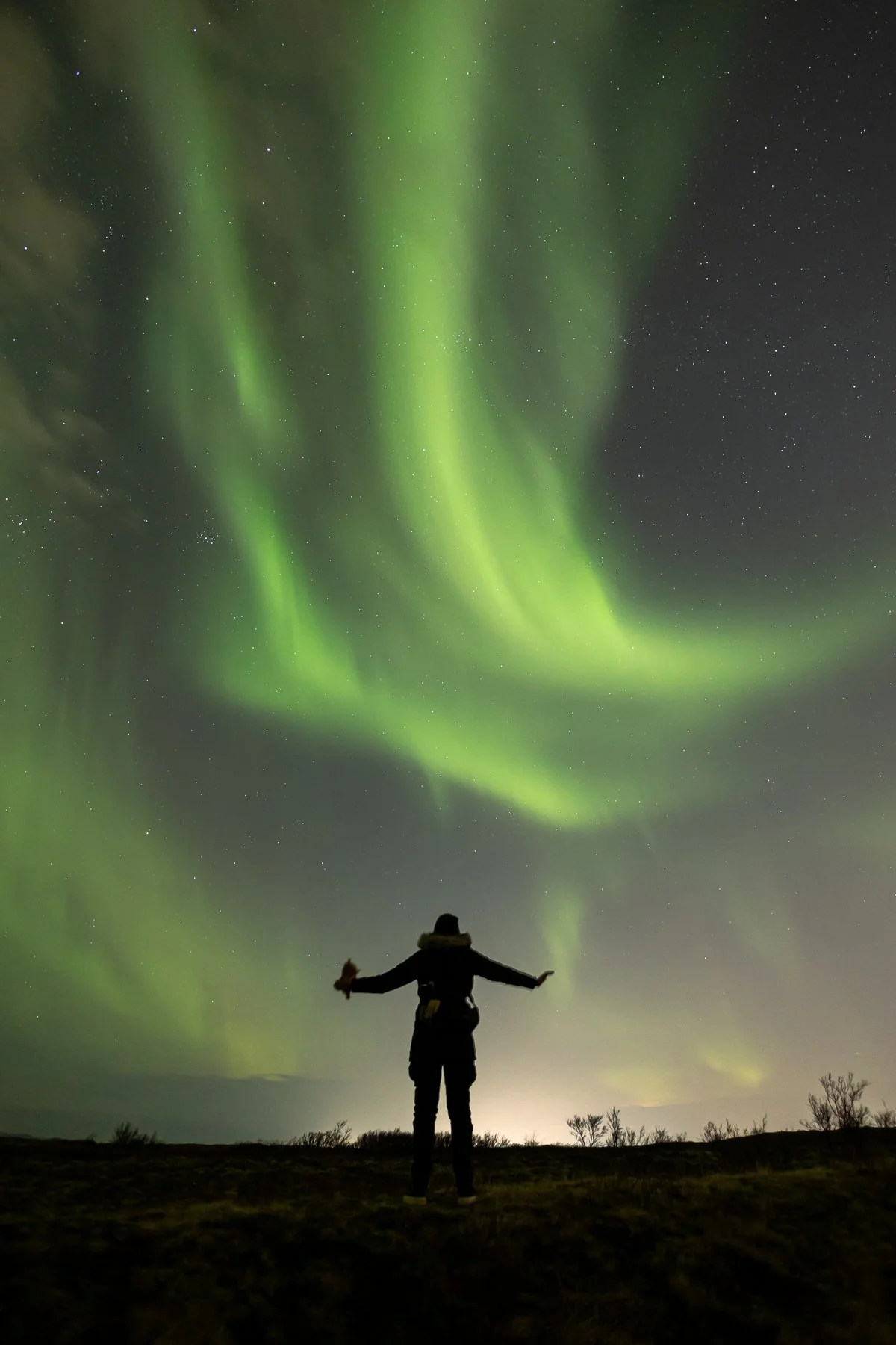 Person standing outdoors at night with arms outstretched, watching the Northern Lights in the sky.