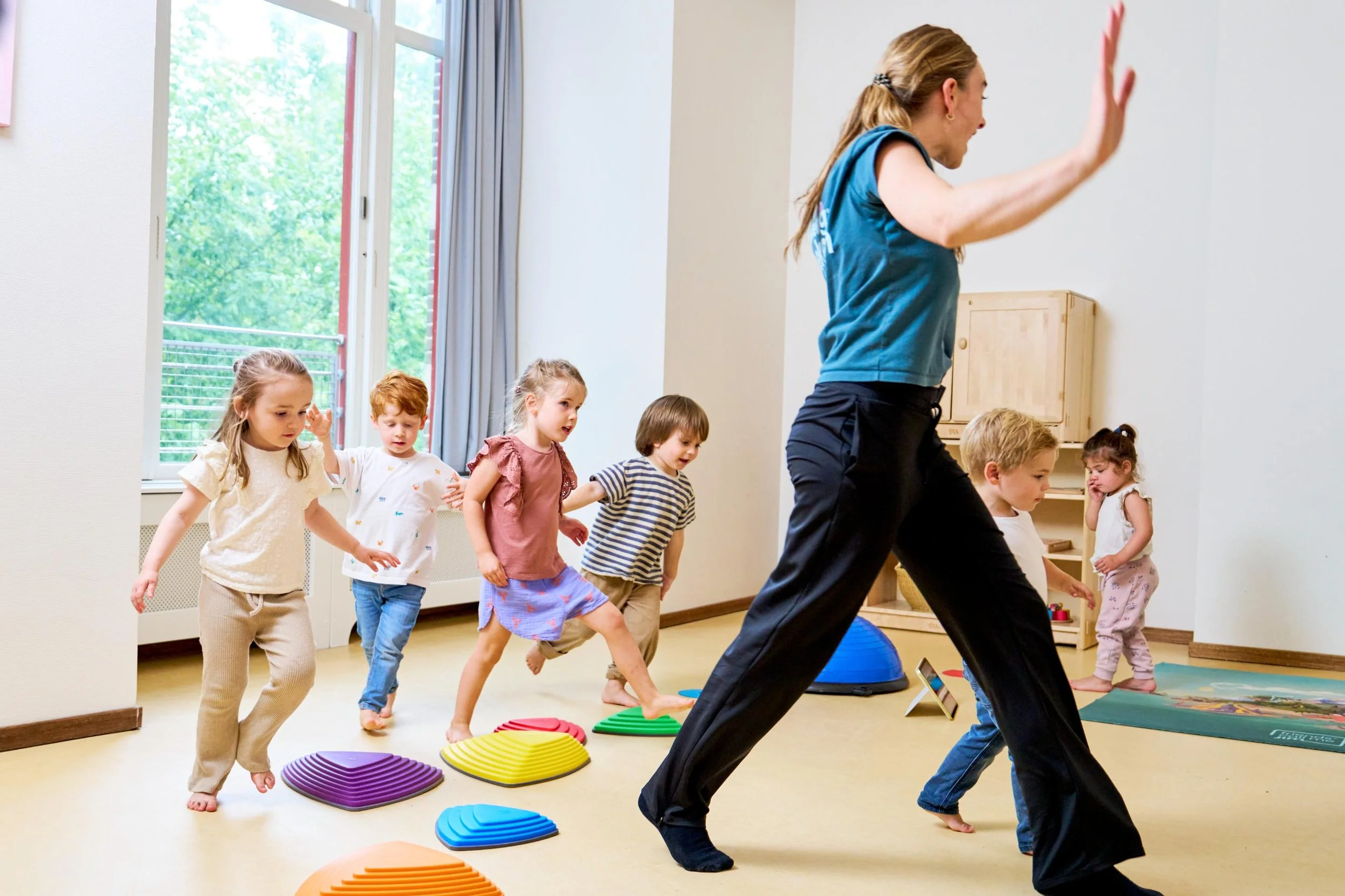 Een vrouw leidt een groep jonge kinderen in een indoors oefening of spel, waarbij ze over kleurrijke opstapjes lopen.