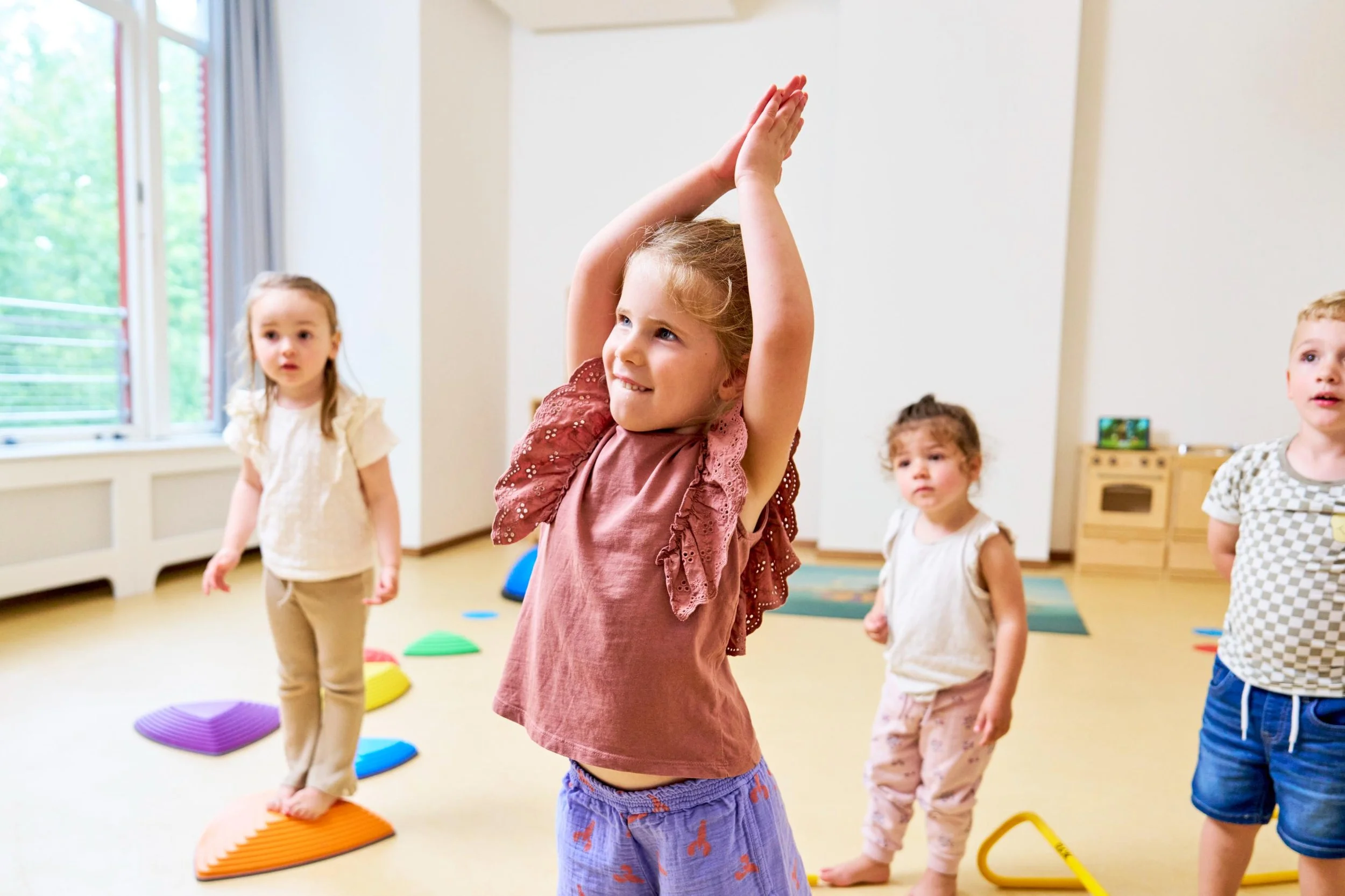 Kinderen spelen en oefenen balans in een lichte kamer met grote ramen, kleurrijke zachte matten en speelgoed.
