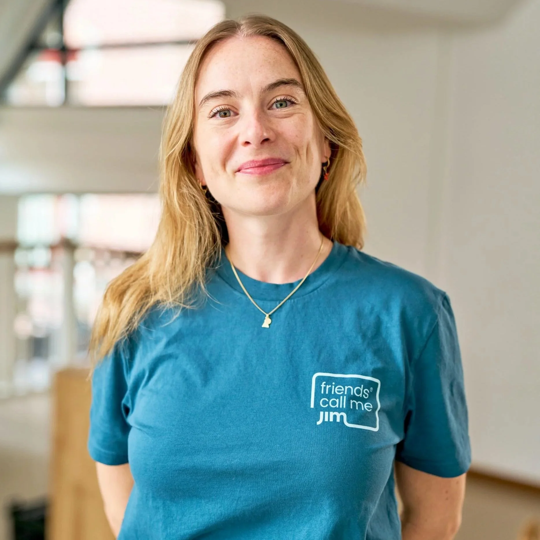 Een vrouw met blond haar draagt een blauwe T-shirt met de tekst 'friends call me JIM' op de voorkant en staat binnen in een gebouw, glimlachend naar de camera.