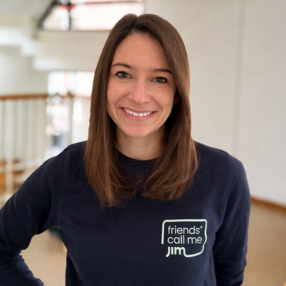 Vrouw met bruin haar glimlachend, zittend in een binnenruimte, draagt een donkerblauw shirt met tekst 'friends' call me JIM' op de borst.