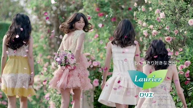 花が咲く庭を背景に、四人の女の子と一人の女性がピンクの花びらを散らしながら歩いている。女性はピンクのドレスを着て、花束を持っている。