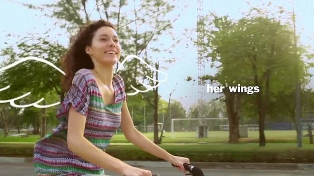 女性が自転車に乗って走っており、背中には白い羽が描かれているように見える。背景は公園の木々と芝生。