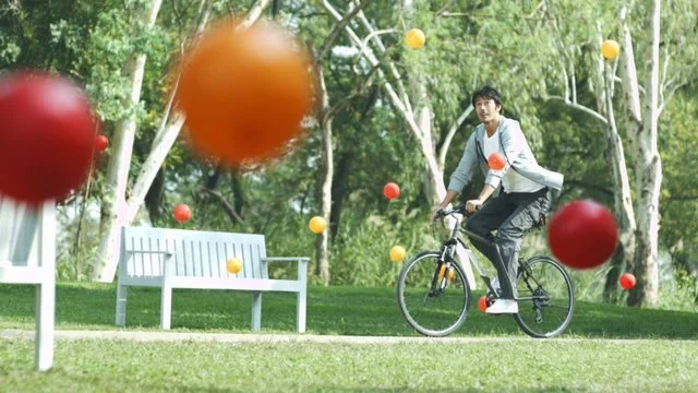 公園で自転車に乗る男性と、色とりどりのボールが空中に浮かんでいる風景