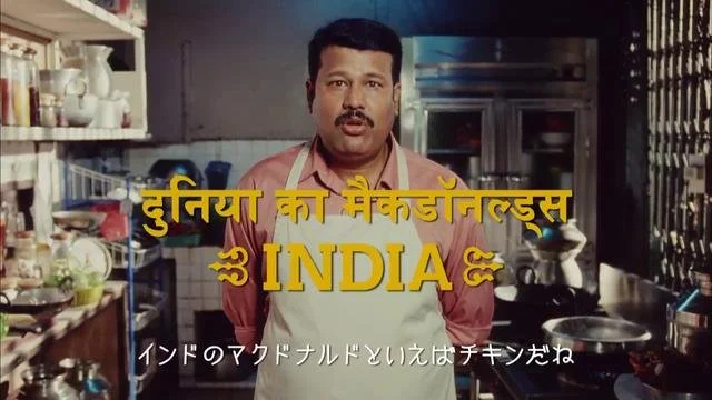厨房にいる男性が中央に立っている。画像には黄色のテキストで『世界のメジャンドレス』と書かれ、その下に『INDIA』と記されている。
