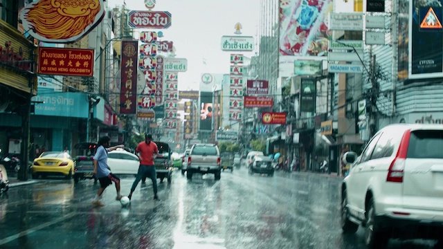 雨の中の都市の繁華街。道路に人々と車が走り、多くの看板とビルが並ぶ。