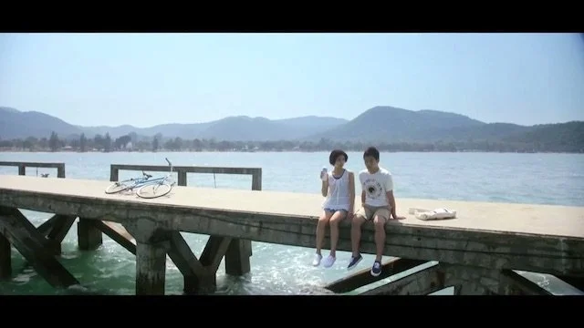 湖のそばの木製の桟橋に座る若い男女二人。水辺に足を浸しながら話す。背景に山と青空が広がる風景。