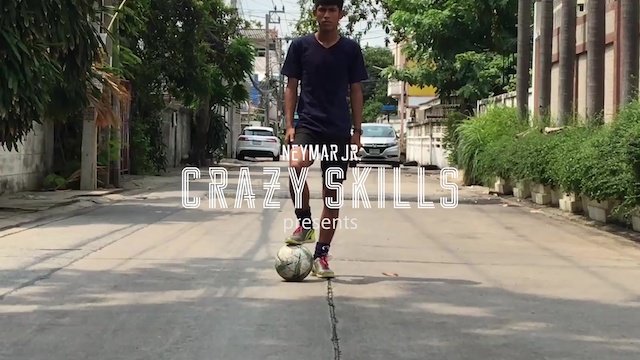 青年が道路でサッカーボールを蹴っている。背景に木々と車、家々が見える。