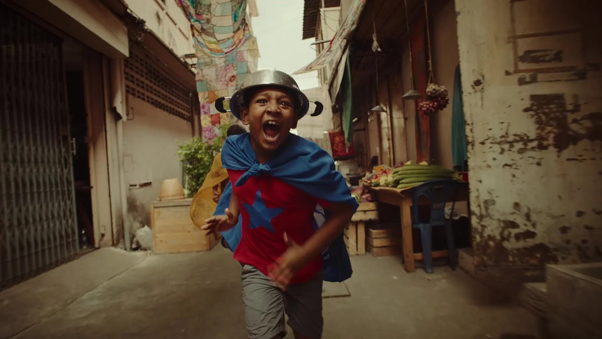 ワイヤーネットのヘルメットをかぶった子供が走っている。背景は市場の通りで、布や野菜が並ぶ屋台や古い建物が見える。