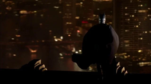 夜景を背景に、バットマンのコスチュームを着た人が建物の窓辺に立っている。