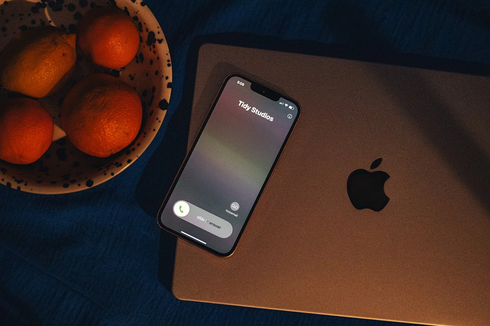 iPhone on closed MacBook with 'Tidy Studios' incoming call, bowl of oranges and lemons on blue fabric surface.