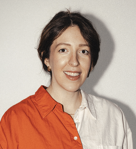 A woman with short brown hair smiling, wearing a white and orange shirt and gold earrings, against a plain light background.