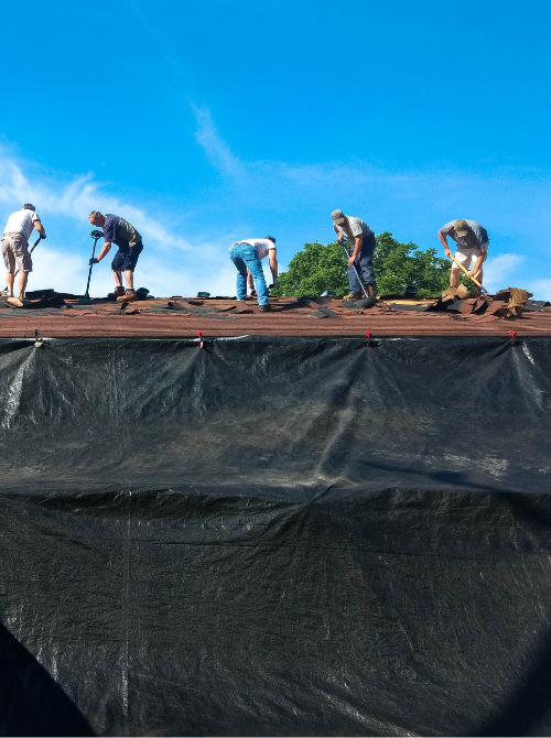 Five workers installing or repairing a roof on a sunny day, with trees and a blue sky in the background.