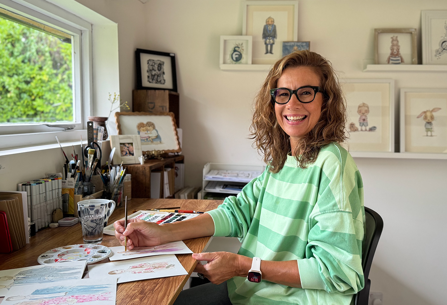 A smiling woman with curly hair, wearing glasses and a green striped sweater, sitting at a wooden desk painting watercolor pictures in a well-lit room with colorful framed artwork on the wall.