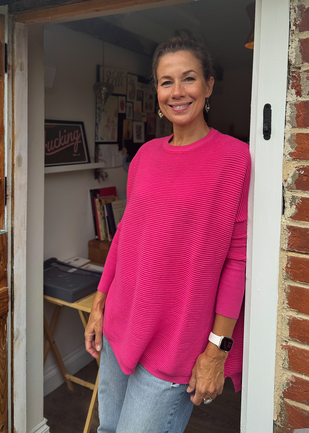 A smiling woman in a pink sweater and jeans standing in a doorway with a brick wall on the right and a decorated interior behind her.