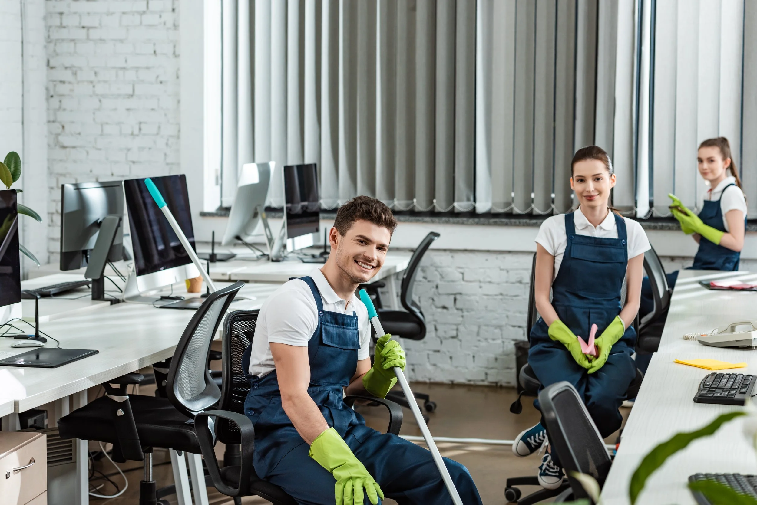 Drei junge Menschen in einem Büro, verkleidet als Reiniger, tragen blaue Overalls und grüne Gummihandschuhe, und posieren lachend für das Foto.