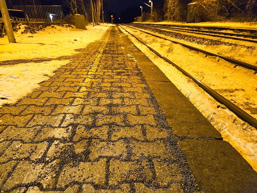 Gepflasterter Gehweg neben Eisenbahnschienen bei Nacht, mit schneebedecktem Umfeld.