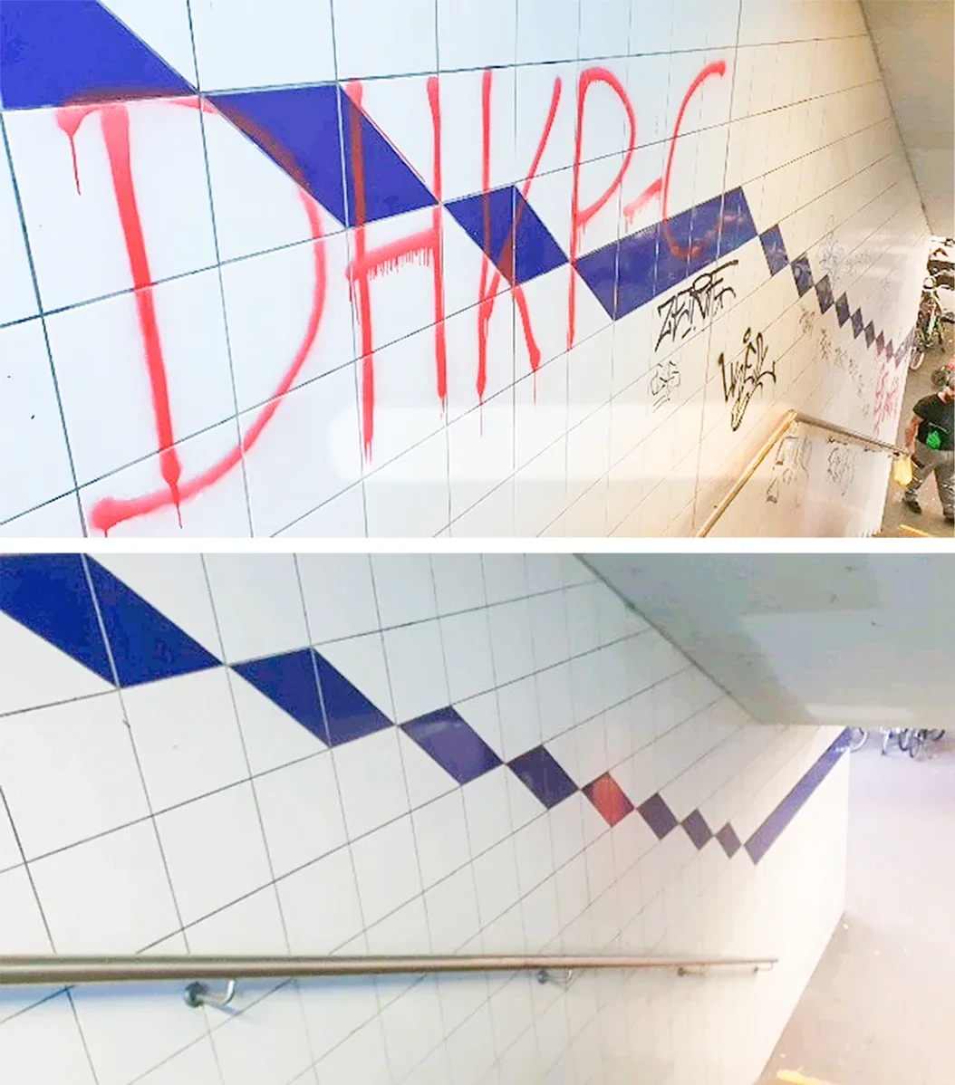 Ein Wandabschnitt in einer U-Bahn-Station mit weißen Fliesen, blauen geometrischen Mustern und Graffiti. Es sind Menschen im unteren rechten Bereich zu sehen.