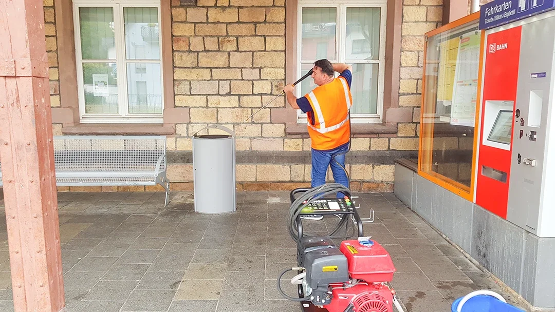 Reinigungskraft mit einem Hochdruckreiniger reinigen eine Mülltonne an einem Bahnhofsgebäude.