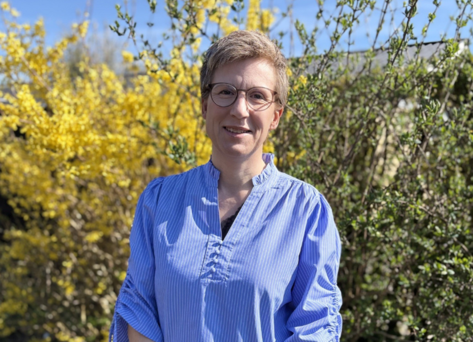 A woman with short blonde hair, glasses, and a blue striped blouse standing outdoors in front of flowering bushes with yellow and white blossoms.