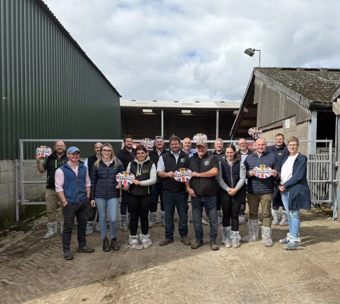 A group of people standing on a farm, holding signs that say 'Back British Farming'. They are wearing shoe covers and are in front of farm buildings.
