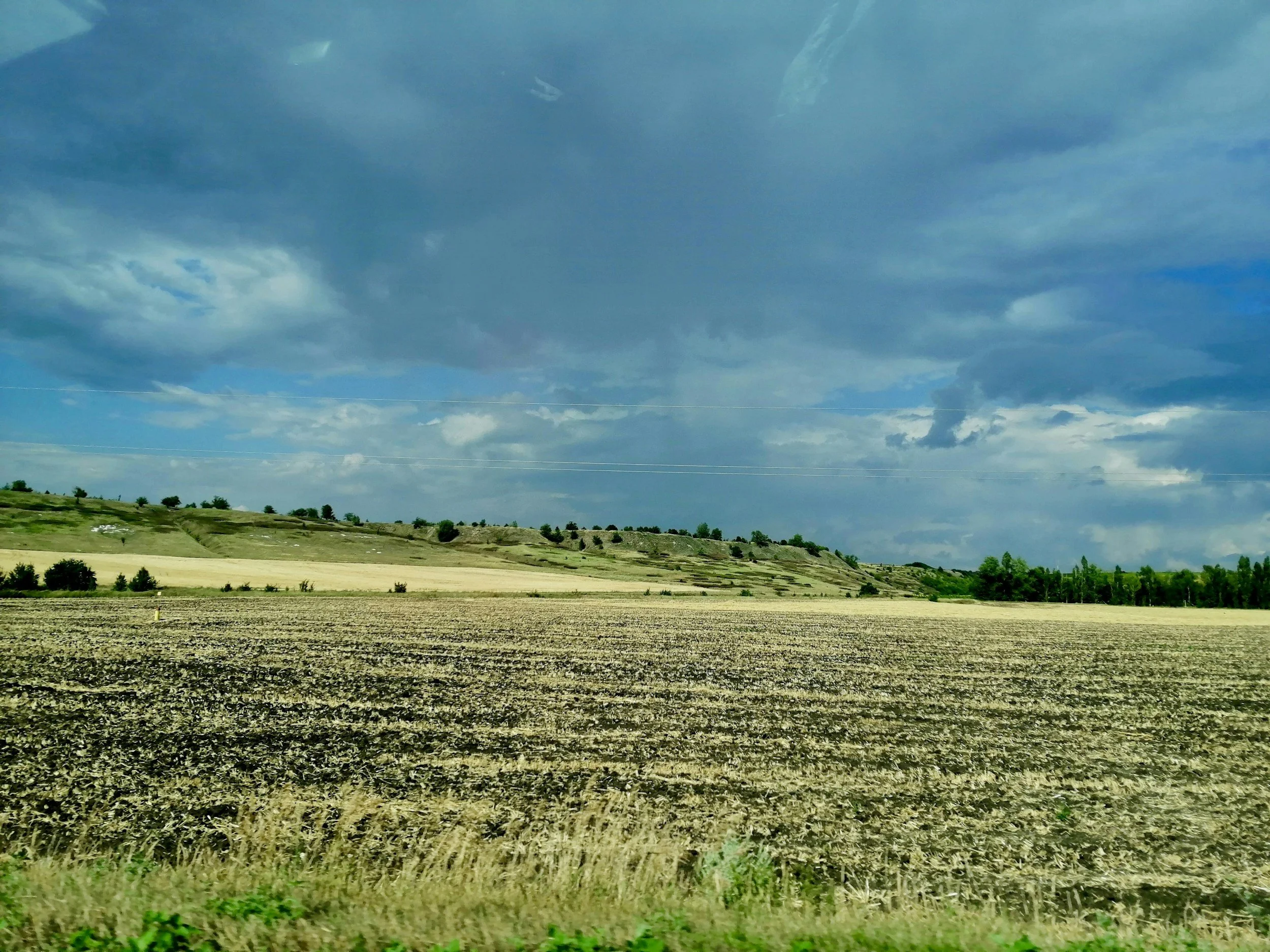 Wide view of a rural landscape with large open farmland, rolling green hills, scattered trees, and a partly cloudy sky.