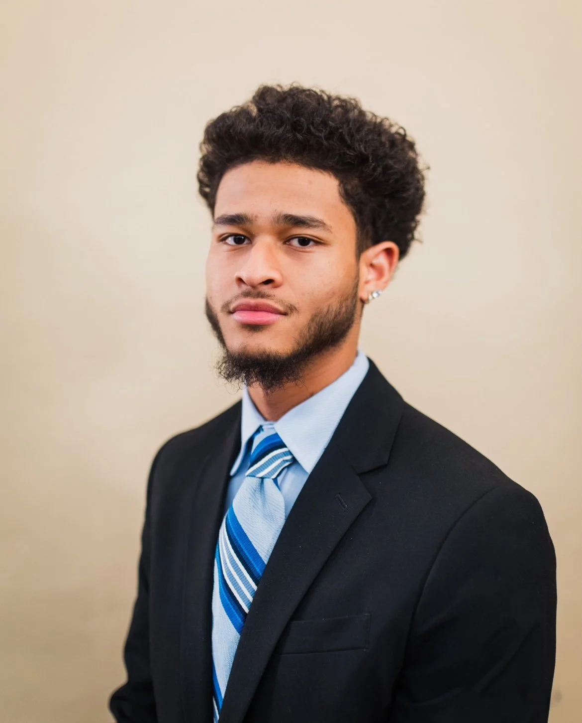 Young man with curly hair and beard wearing a black suit, light blue shirt, and striped tie, standing in front of a plain beige background.